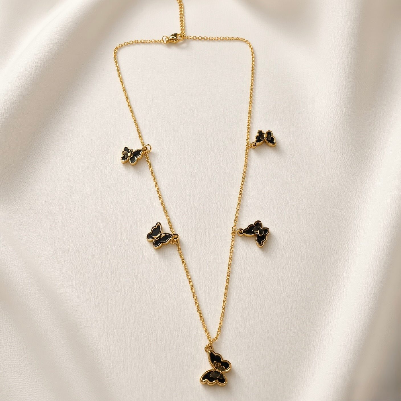Papillon Noir Black Enamel Butterfly Charm Necklace - full length flat lay on ivory silk showing complete necklace laid out: fine gold oval-link cable chain with five graduated black enamel butterfly charms at asymmetric intervals showing size graduation from small upper butterflies to large central drop, lobster clasp at top, deep black enamel fill with warm gold border on each butterfly charm clearly visible