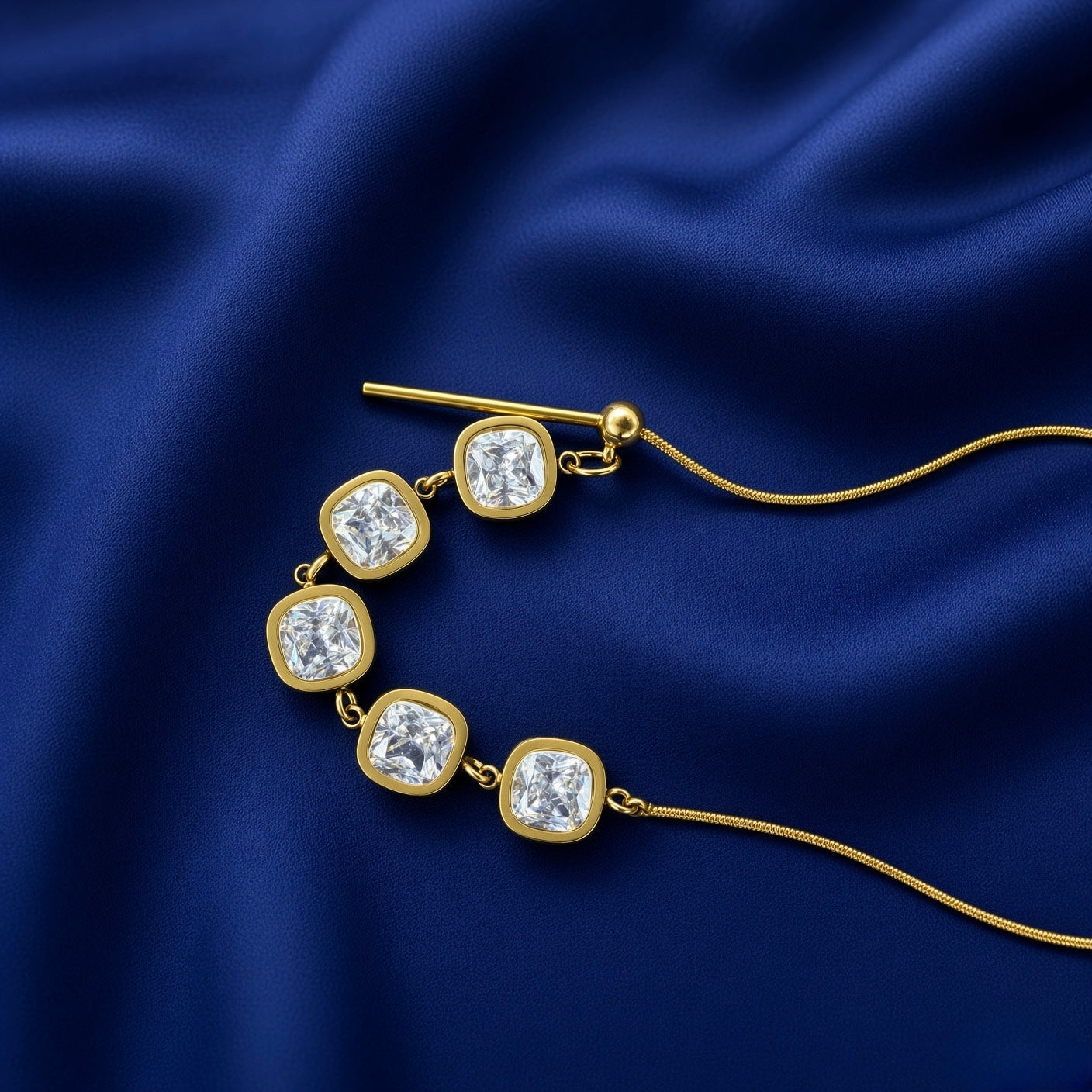 Lumière Cinq Cushion Crystal Convertible Snake Chain Necklace - macro close-up on deep navy velvet showing five-stone arc detail: four cushion-cut diamond-clear crystal zirconia in smooth continuous gold bezels linked by gold jump rings, gold bar-and-ball toggle mechanism at left with gold ball stop and bar extending downward, flat-link snake chain visible at right