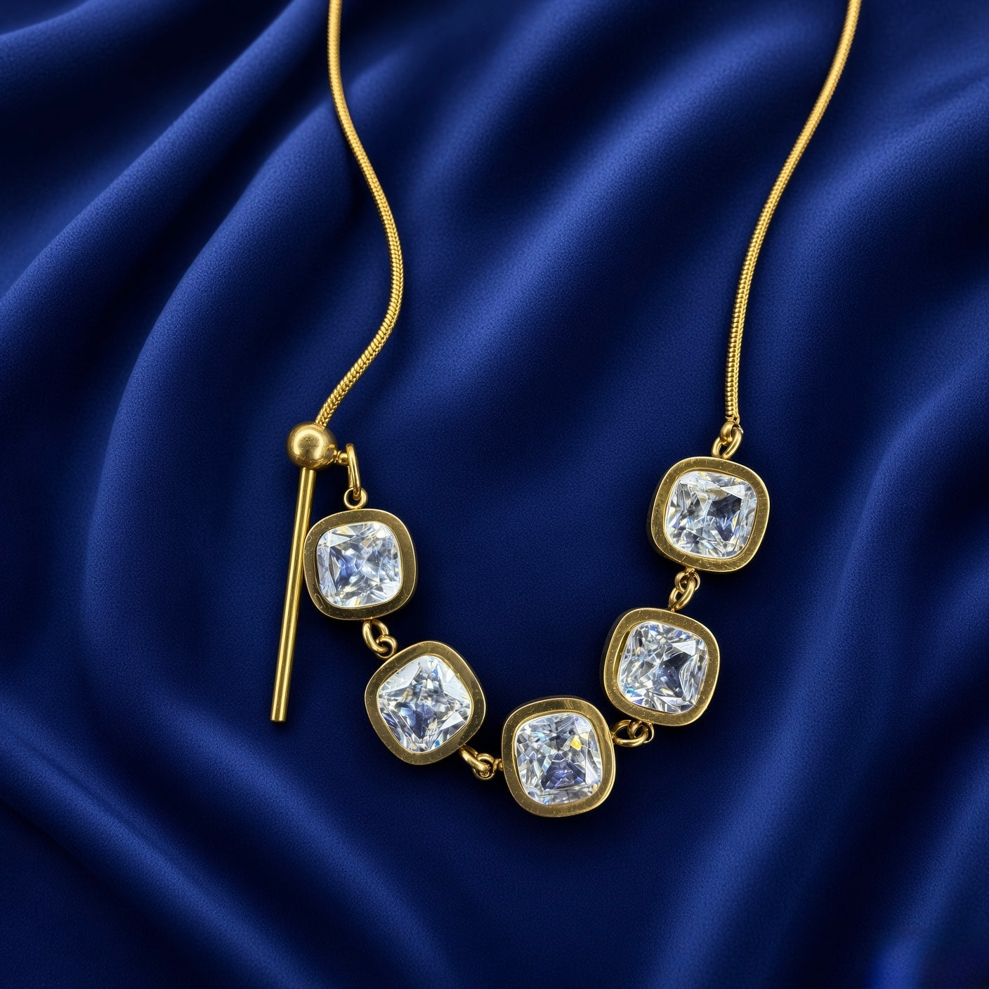 Lumière Cinq Cushion Crystal Convertible Snake Chain Necklace - three-quarter close-up on deep navy velvet showing five cushion-cut crystal zirconia station arc: five generous cushion-cut diamond-clear crystals in smooth gold bezels linked in continuous arc, gold bar-and-ball toggle at left, flat-link snake chain on both sides, pure white diamond-like brilliance of cushion crystals clearly visible