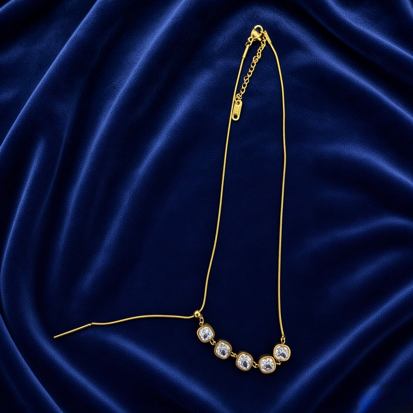 Lumière Cinq Cushion Crystal Convertible Snake Chain Necklace - full flat lay on deep navy velvet in lariat Y-drop mode showing convertible silhouette: flat-link gold snake chain with five cushion-cut crystal zirconia arc pulled into vertical Y-drop by gold bar-and-ball toggle mechanism, stones cascading downward in dramatic lariat configuration, lobster clasp and extender chain at top right