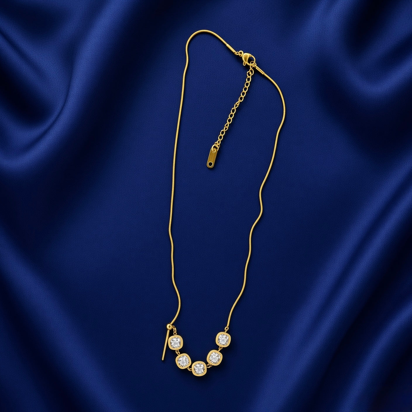 Lumière Cinq Cushion Crystal Convertible Snake Chain Necklace - full flat lay on deep navy velvet in station mode from slightly different angle showing complete necklace: flat-link gold snake chain with five cushion-cut diamond-clear crystal zirconia in smooth gold bezels arcing symmetrically across front in classic station configuration, gold bar-and-ball toggle at left, lobster clasp and branded extender chain tag at top right