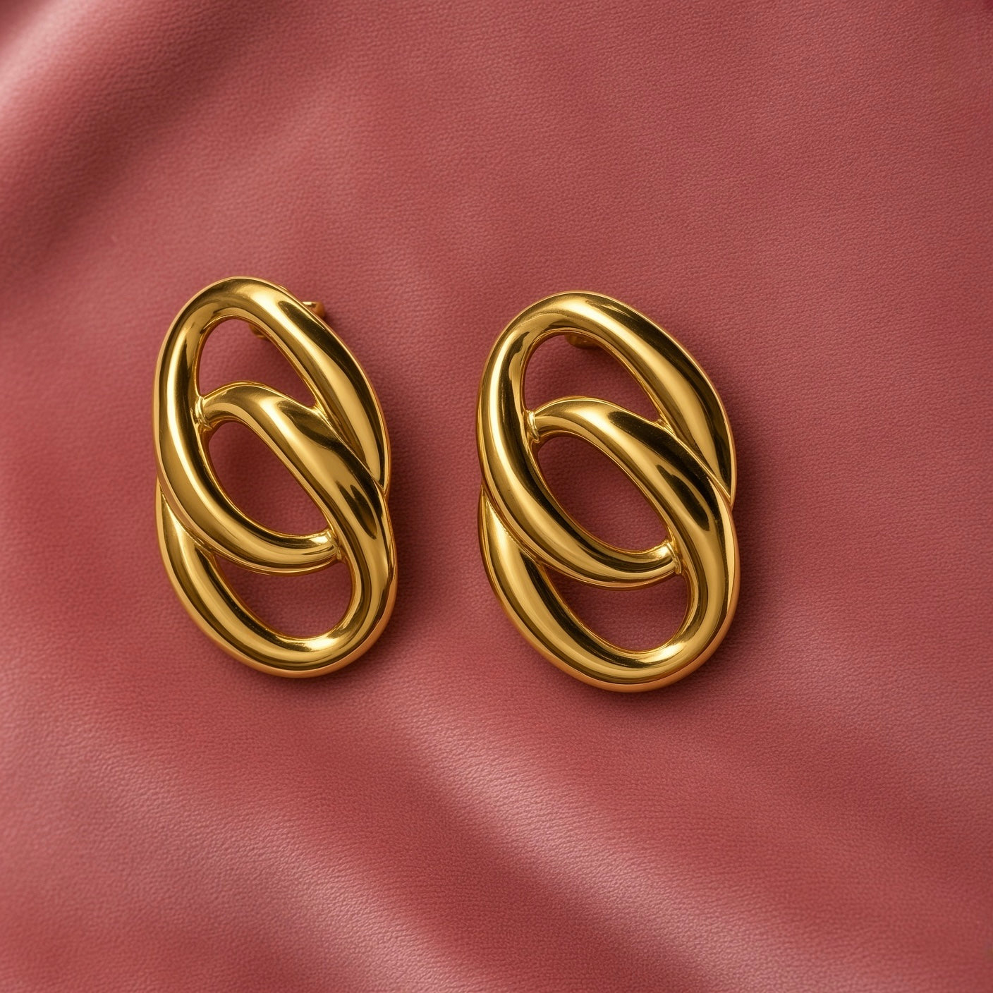 Gold Double Oval Interlocked Link Stud Earrings - pair face-on on dusty pink satin showing complete earrings from slightly wider angle: vertical oval silhouette with large outer oval and smaller inner oval interlocked, high mirror-polish warm gold surface reflecting dusty pink satin background, bold statement scale and three-dimensional chain-link construction clearly visible