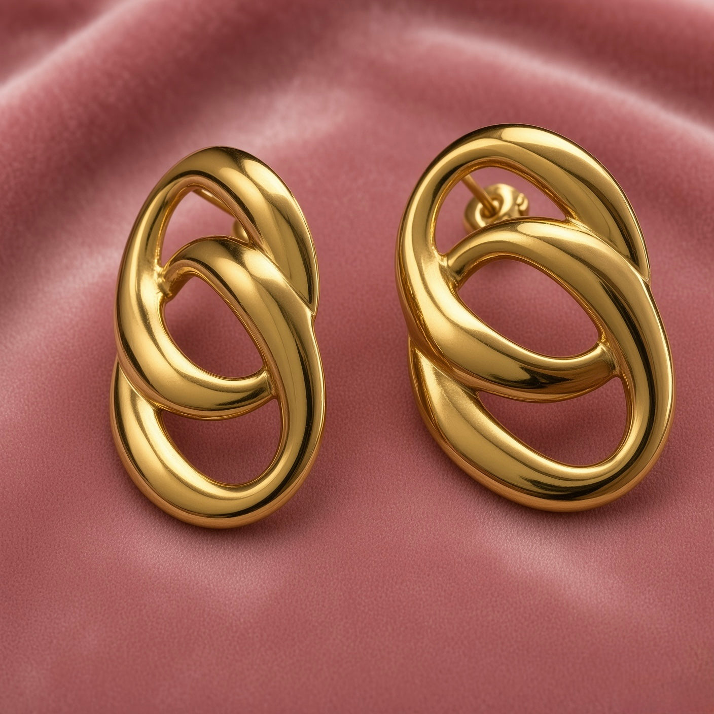 Gold Double Oval Interlocked Link Stud Earrings - pair elevated three-quarter close-up on dusty pink satin showing full three-dimensional depth of double oval interlock: large outer oval tube and smaller inner oval tube passing through each other, thick rounded tube walls in high mirror-polish gold, warm gold highlights and shadows of interlocked oval composition, straight post with butterfly clutch back visible on right earring