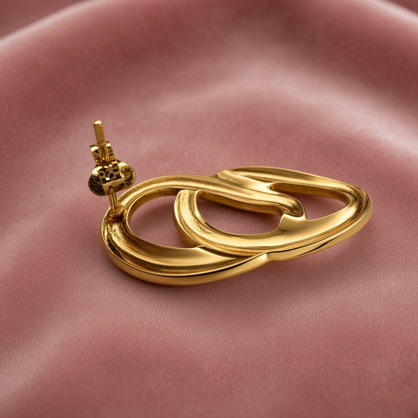 Gold Double Oval Interlocked Link Stud Earrings - side view on dusty pink satin showing single earring three-dimensional construction: large outer oval tube and smaller inner oval tube genuinely interlocked in full 3D, thick rounded tube walls in mirror-polish gold, straight gold post with butterfly friction clutch back visible at top rear