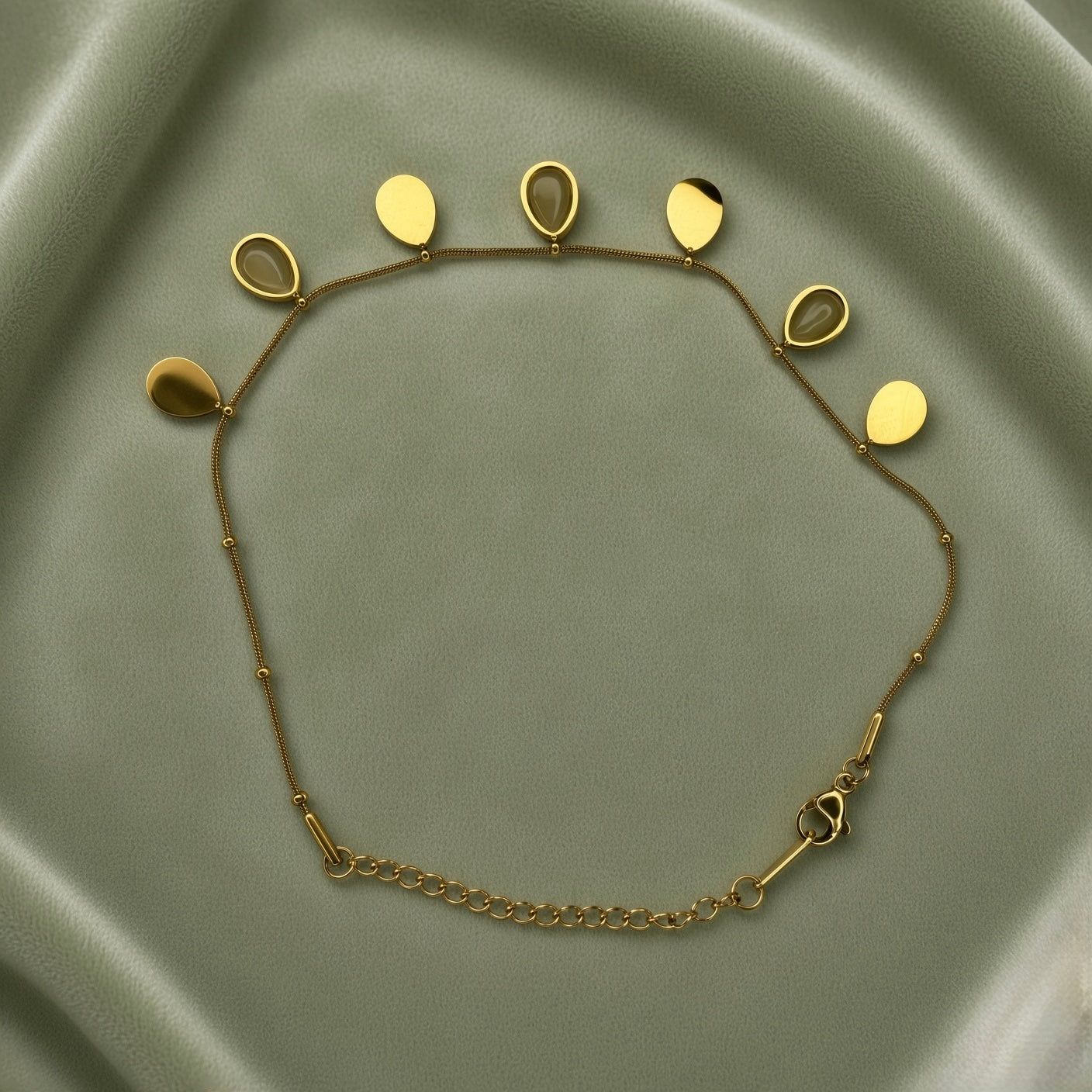 Gold Teardrop Charm Satellite Chain Anklet - full flat lay on sage green satin showing complete anklet from slightly closer angle: fine satellite chain with seven alternating teardrop charms showing solid gold teardrops and bezel-set nude cabochon teardrops in alternating composition, lobster clasp and curb-link extender chain at bottom right, gold ball stations along chain clearly visible