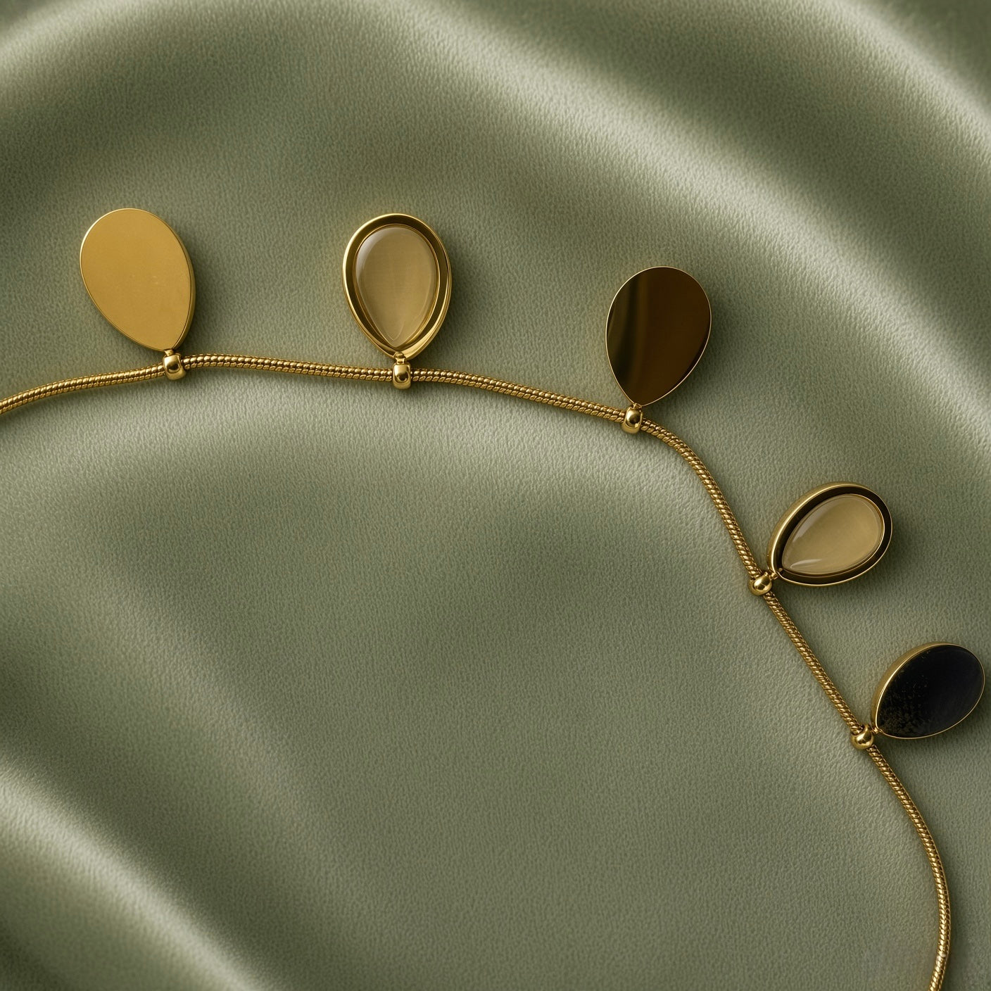 Gold Teardrop Charm Satellite Chain Anklet - macro close-up on sage green satin showing four teardrop charms in detail: solid mirror-polish warm gold flat teardrop charm (left), open gold oval bezel frame with smooth nude taupe champagne cabochon stone teardrop charm (centre left), solid mirror-polish gold teardrop charm (centre right), open gold bezel nude cabochon teardrop charm (right), fine satellite chain with gold ball stations below all charms