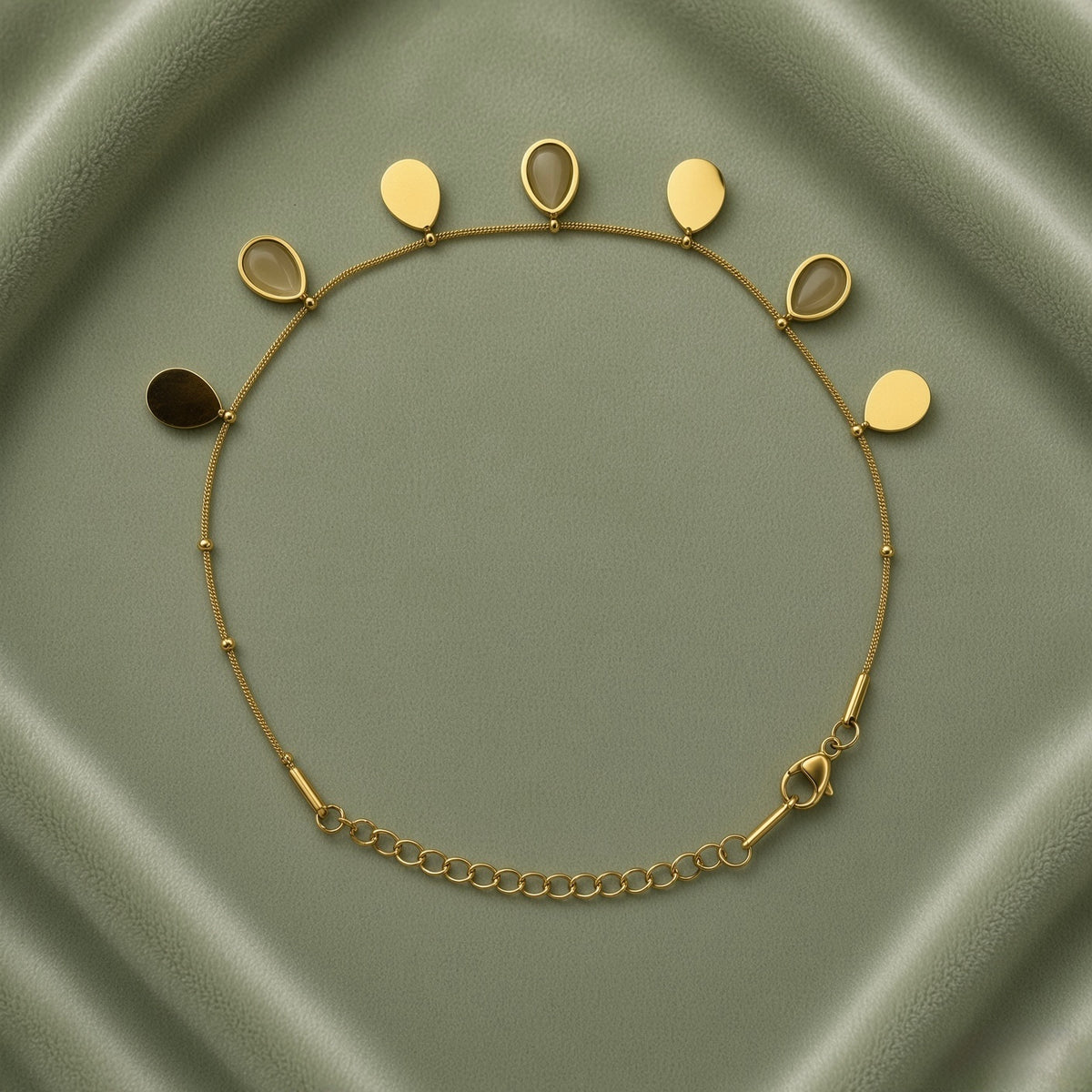 Gold Teardrop Charm Satellite Chain Anklet - full flat lay on sage green satin tray showing complete anklet in circular layout: fine gold satellite chain with evenly spaced gold ball stations carrying seven alternating teardrop charms along upper arc, solid mirror-polish gold flat teardrop charms alternating with open gold bezel nude cabochon teardrop charms, lobster clasp and curb-link extender chain at bottom right