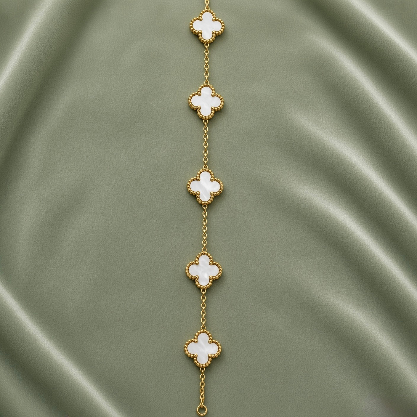 White Clover Station Bracelet Gold Milgrain - full length flat lay on sage green satin showing five clover motifs in vertical arrangement: five white mother-of-pearl four-leaf clover quatrefoil motifs with continuous milgrain gold bead borders macro detail, smooth luminous white mother-of-pearl fill and precise small gold ball milgrain border on each clover clearly visible, fine gold cable link chain connecting all five motifs