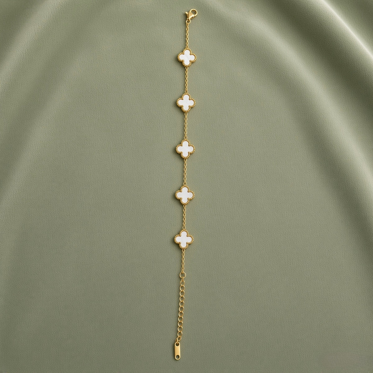 White Clover Station Bracelet Gold Milgrain - full length flat lay on sage green satin showing complete bracelet laid straight: five white mother-of-pearl four-leaf clover motifs with continuous milgrain gold bead borders evenly spaced on fine gold cable link chain, lobster clasp at top, curb-link extender chain and gold barrel tag at bottom