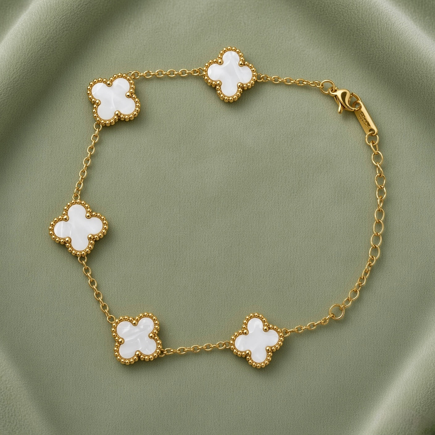 White Clover Station Bracelet Gold Milgrain - circular flat lay on sage green satin showing complete bracelet in loop: five white mother-of-pearl clover motifs with milgrain gold bead borders on fine gold cable link chain, lobster clasp visible at right, white mother-of-pearl fill and milgrain bead border detail clearly visible on all five clover motifs