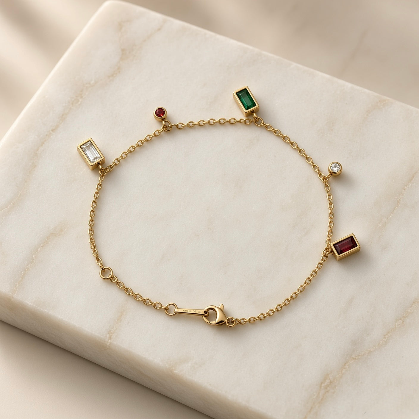 Multicolour Gemstone Bezel Charm Bracelet Gold - circular flat lay on cream satin: five bezel-set dangle charms on fine gold cable chain — white baguette left, ruby red round and emerald green baguette top, diamond-clear white round right, deep ruby red baguette bottom right, lobster clasp at bottom centre