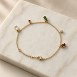 Multicolour Gemstone Bezel Charm Bracelet Gold - circular flat lay on cream satin: five bezel-set dangle charms on fine gold cable chain — white baguette left, ruby red round and emerald green baguette top, diamond-clear white round right, deep ruby red baguette bottom right, lobster clasp at bottom centre
