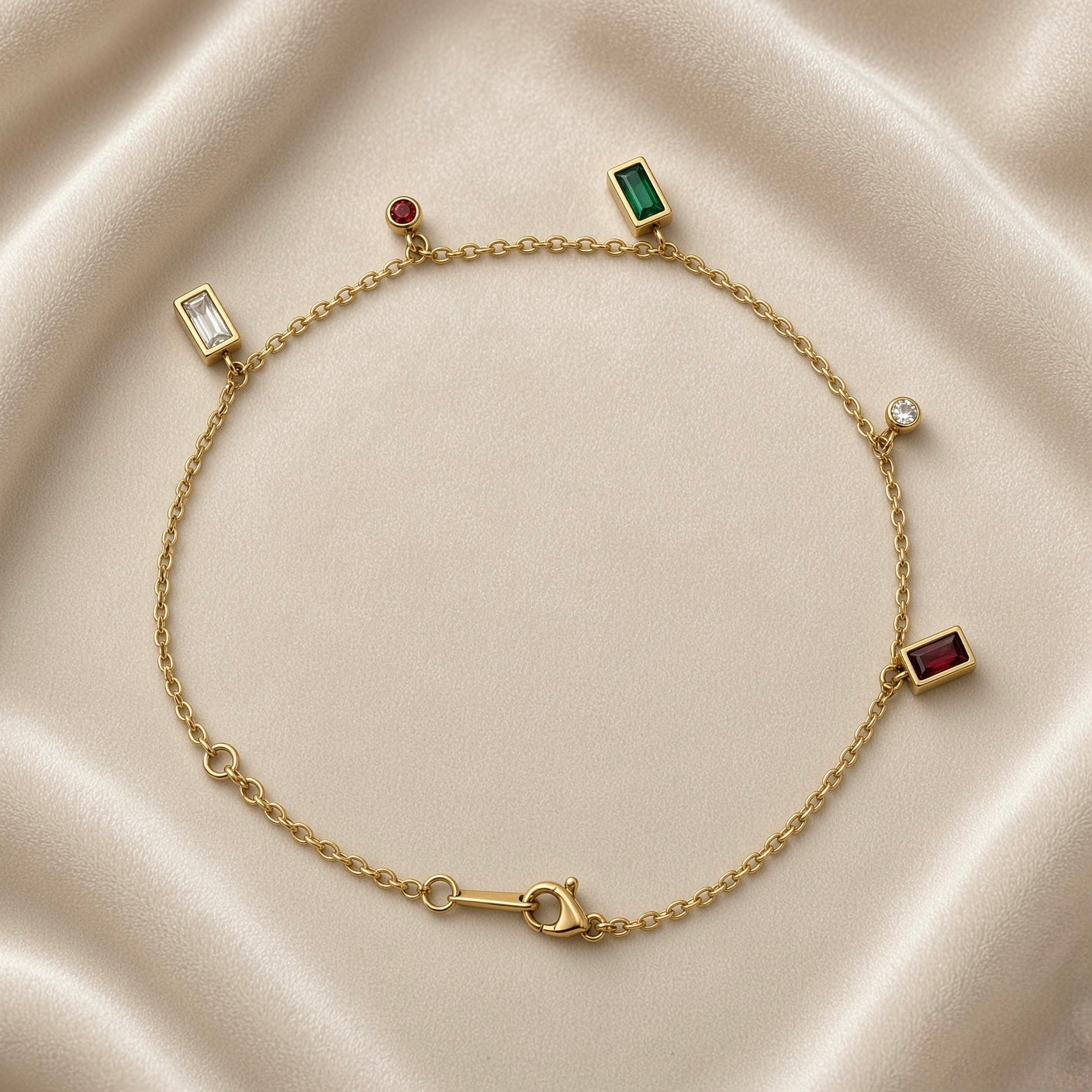 Multicolour Gemstone Bezel Charm Bracelet Gold - circular flat lay on white marble: fine gold cable chain with five bezel charms — white baguette bezel left, red round bezel and emerald green baguette bezel top, white round bezel right, ruby red baguette bezel bottom right, lobster clasp with engraved gold bar tag at bottom