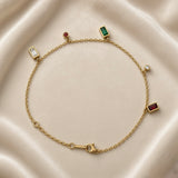 Multicolour Gemstone Bezel Charm Bracelet Gold - circular flat lay on white marble: fine gold cable chain with five bezel charms — white baguette bezel left, red round bezel and emerald green baguette bezel top, white round bezel right, ruby red baguette bezel bottom right, lobster clasp with engraved gold bar tag at bottom