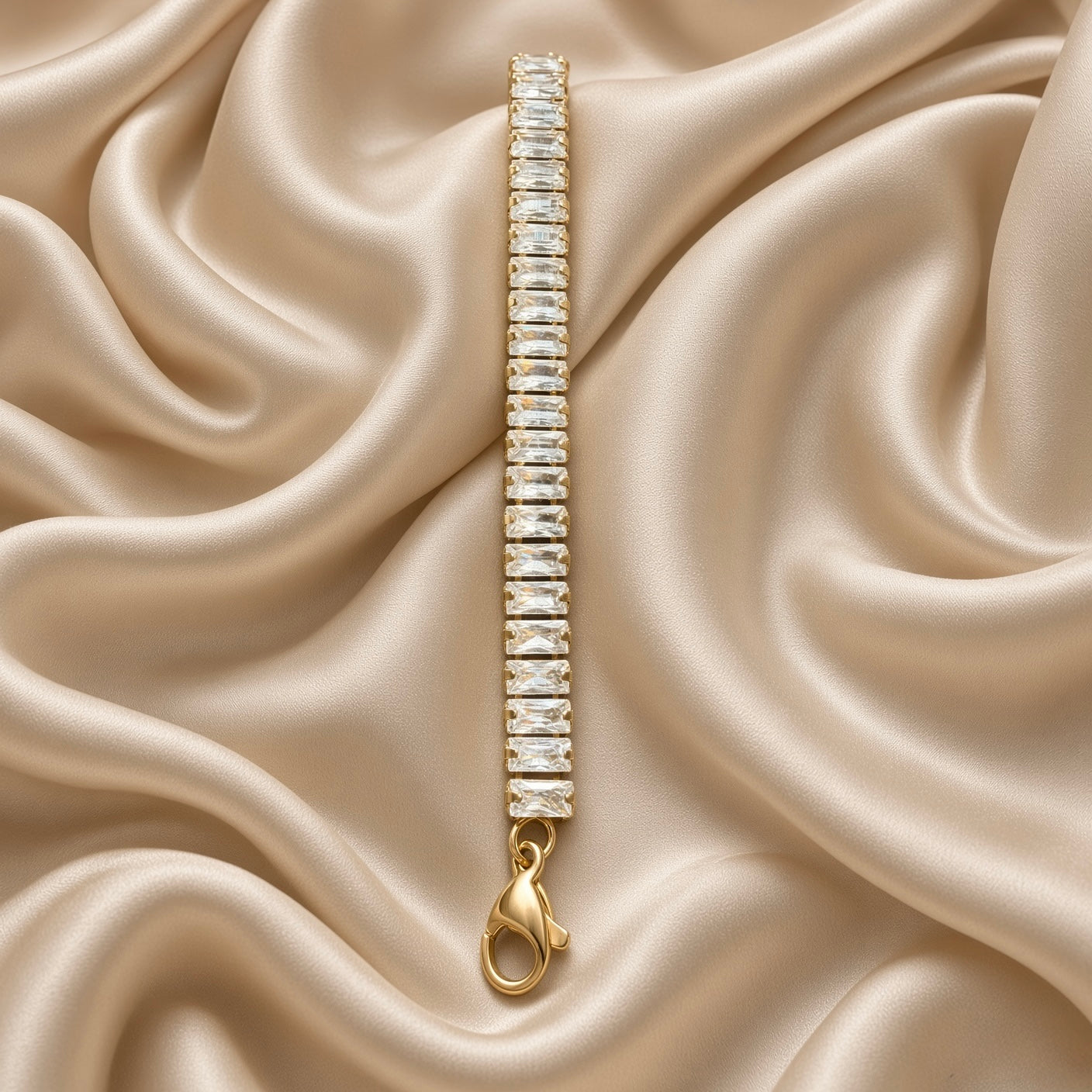 Baguette Crystal Tennis Bracelet Gold - diagonal flat lay on cream satin showing full baguette channel band detail: step-cut white crystal zirconia stones in continuous channel setting, lobster clasp with gold tag at bottom