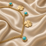Gold Clover and Turquoise Charm Bracelet Curb Chain - macro detail on cream satin: bold gold curb chain showing flat mirror-polish gold four-leaf clover charm with four heart-shaped petals and engraved centre, round turquoise cabochon bezel charm with vivid sky blue-green stone and natural matrix markings in gold bezel