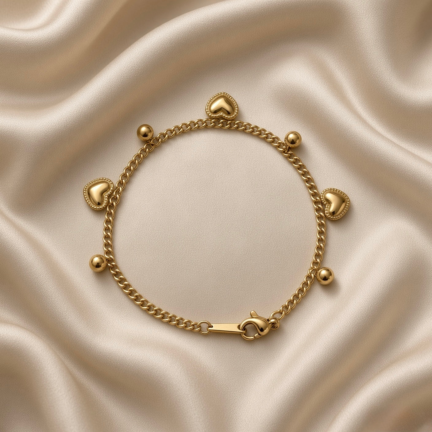 Gold Heart and Ball Charm Bracelet Milgrain Curb Chain - vertical full-length flat lay on cream satin: bold gold curb chain with six alternating dangle charms, three puffy milgrain-border gold heart charms and three smooth round gold ball charms, engraved gold tab at top, lobster clasp at base