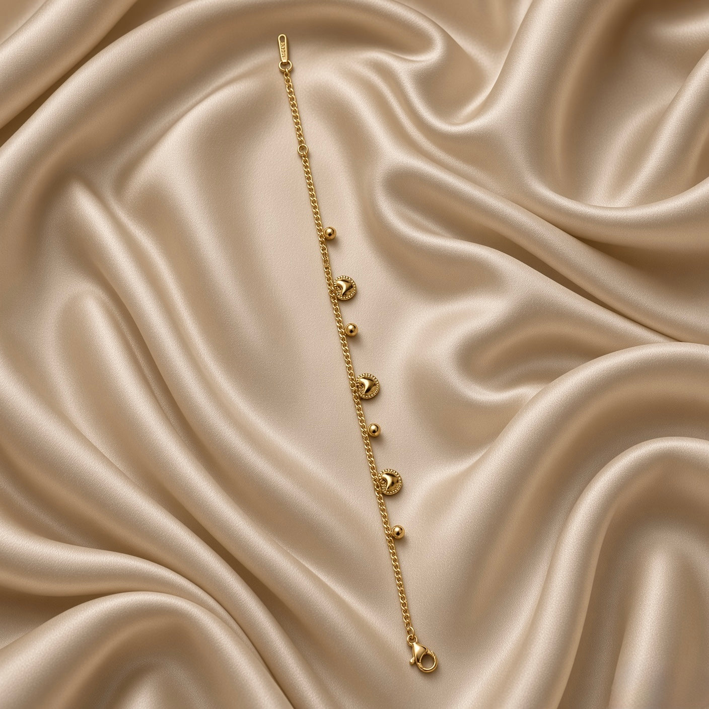 Gold Heart and Ball Charm Bracelet Milgrain Curb Chain - circular flat lay on cream satin showing full bracelet loop: bold gold curb chain with three puffy milgrain gold heart charms and three smooth round gold ball charms alternating, lobster clasp with engraved gold bar tag at bottom centre