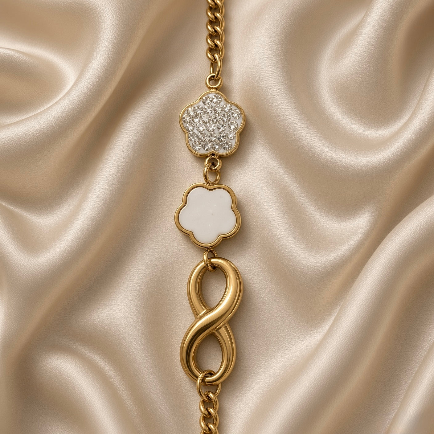 Gold Infinity Clover Crystal Flower Charm Bracelet Curb Chain - vertical full-length flat lay on cream satin: bold gold curb chain showing three cascading charms — white crystal pave five-petal flower top, white enamel clover centre, bold tubular gold infinity bottom, lobster clasp at base