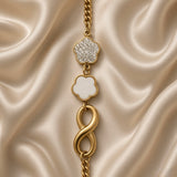 Gold Infinity Clover Crystal Flower Charm Bracelet Curb Chain - vertical full-length flat lay on cream satin: bold gold curb chain showing three cascading charms — white crystal pave five-petal flower top, white enamel clover centre, bold tubular gold infinity bottom, lobster clasp at base