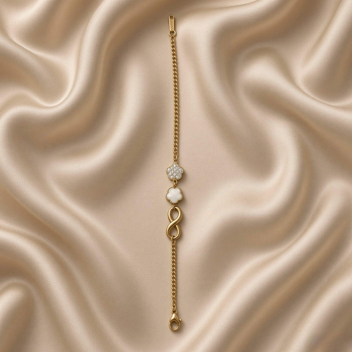 Gold Infinity Clover Crystal Flower Charm Bracelet Curb Chain - circular flat lay on cream satin: bold gold curb chain with three cascading charms at top — white crystal pave flower, white enamel quatrefoil clover, bold 3D gold infinity, flat gold ID bar tag and lobster clasp at bottom