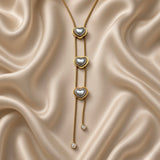 Pearl Heart Lariat Necklace Triple Station Crystal Drop Gold - close detail on cream satin: three bezel-set pearl heart stations in descending vertical cascade on double-strand box chain drop, two round white crystal zirconia bezel drop terminators at base, pearl hearts showing silver-lustre in directional light, warm 18K gold heart bezels