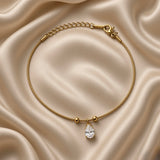 Pear Crystal Solitaire Drop Bracelet Snake Chain Gold - circular flat lay on cream satin: full bracelet loop showing round ribbed gold snake chain, single pear crystal solitaire drop with four-prong gold claw setting hanging from two gold ball satellites at bottom centre, lobster clasp with engraved gold bar tag at top right, curb extender chain