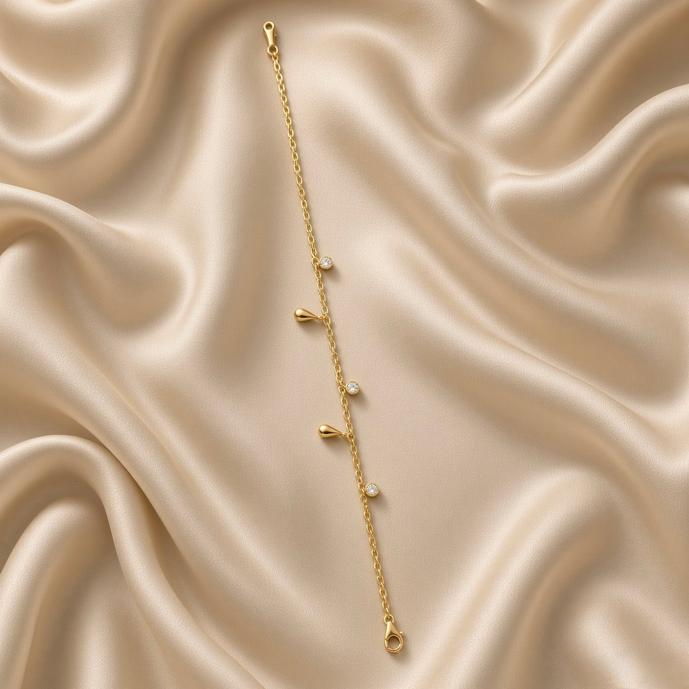 Gold Raindrop and Crystal Bezel Station Bracelet - circular flat lay on cream satin showing full bracelet loop: five alternating charms visible — two solid mirror-polish gold raindrop charms and three round white crystal zirconia bezel drop charms on fine gold cable chain, lobster clasp with gold barrel connector at bottom