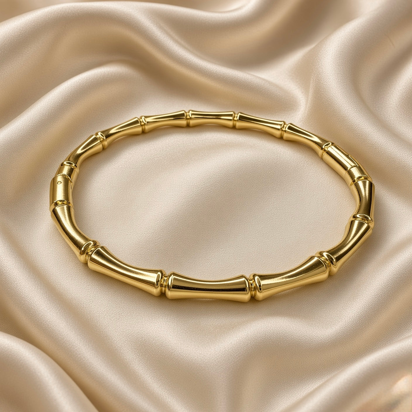 Gold Bamboo Bangle Bracelet Mirror Polish Sculptural - overhead flat lay on cream satin: top-down view of full circular bangle showing all bamboo stalk segments and double-ring joint nodes in mirror-polish warm gold, even warm gold tone across all tube sections, cream satin background