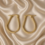 Gold Teardrop Hoop Earrings Tube Round Section Mirror Polish Latch Back Clasp - hero overhead flat lay on cream satin: pair of teardrop tube hoop earrings side by side showing elongated pear teardrop silhouette with wider fuller rounded base tapering to narrower top with latch-back clasp and barrel hinge, round-section circular-profile warm 18K gold tube body, mirror-polish warm gold surface, cream satin background