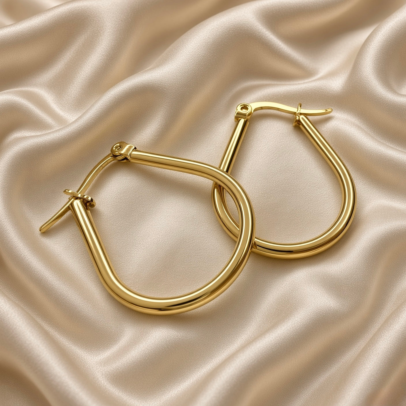 Gold Teardrop Hoop Earrings Tube Round Section Mirror Polish Latch Back Clasp - angled pair flat lay on cream satin: pair of teardrop hoops shown at angle with hoops overlapping, showing three-dimensional round tubular cross-section of hoop body, mirror-polish warm gold surface from multiple angles, latch-back clasp with straight post open at left earring, cream satin background