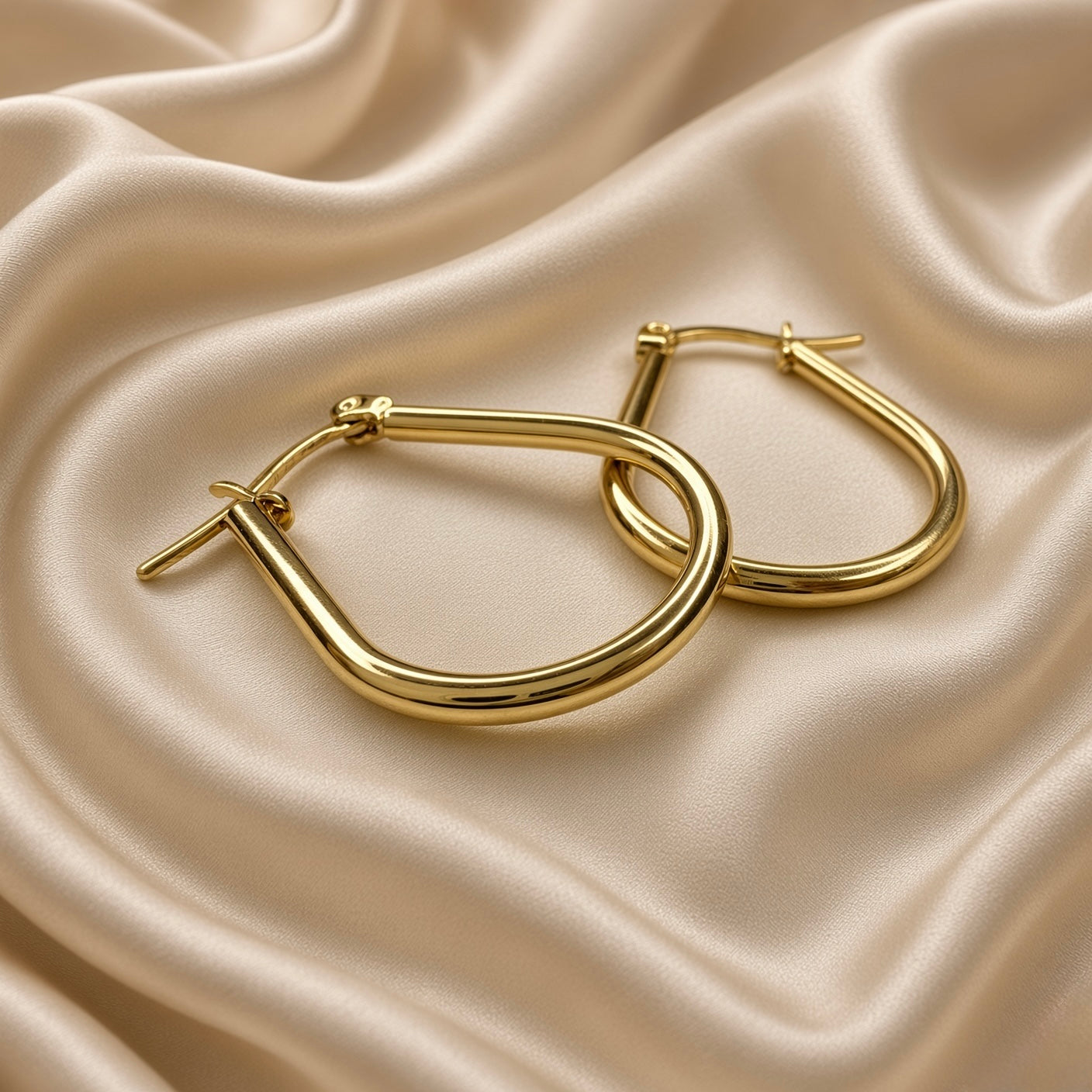 Gold Teardrop Hoop Earrings Tube Round Section Mirror Polish Latch Back Clasp - single earring hero detail on cream satin: single teardrop tube hoop earring showing complete elongated pear teardrop silhouette from rounded base through full tubular round-section hoop body to hinged latch-back clasp at top with barrel hinge detail and straight post visible, mirror-polish warm 18K gold surface catching light as rolling warm gold specular highlight across tube body, cream satin background