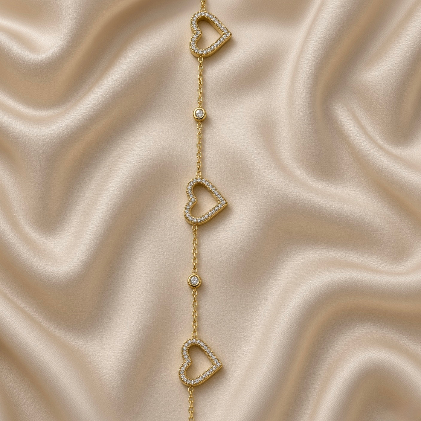 Gold Open Pavé Heart Charm Bracelet Bezel Crystal Station Chain - overhead flat lay on cream satin: full bracelet in circular form showing three open micro-pavé white crystal zirconia heart charms and two round bezel crystal stations on fine warm 18K gold cable chain, lobster clasp at top, cream satin background