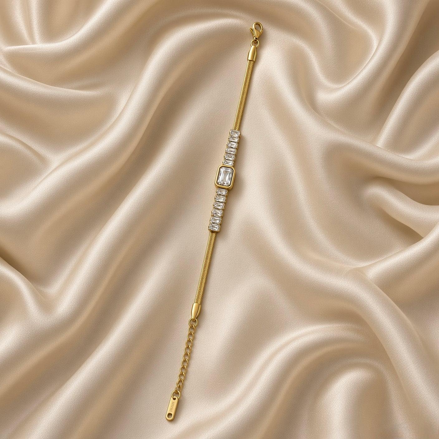 Gold Herringbone Chain Bracelet Baguette Crystal Station Emerald Bezel Centre - angled flat lay on cream satin: three-quarter view of bracelet showing herringbone chain texture detail and baguette crystal channel row with emerald bezel centre, warm gold V-pattern herringbone weave surface visible, white crystal baguette station row and emerald cut bezel focal in warm gold, cream satin background