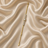 Gold Herringbone Chain Bracelet Baguette Crystal Station Emerald Bezel Centre - angled flat lay on cream satin: three-quarter view of bracelet showing herringbone chain texture detail and baguette crystal channel row with emerald bezel centre, warm gold V-pattern herringbone weave surface visible, white crystal baguette station row and emerald cut bezel focal in warm gold, cream satin background