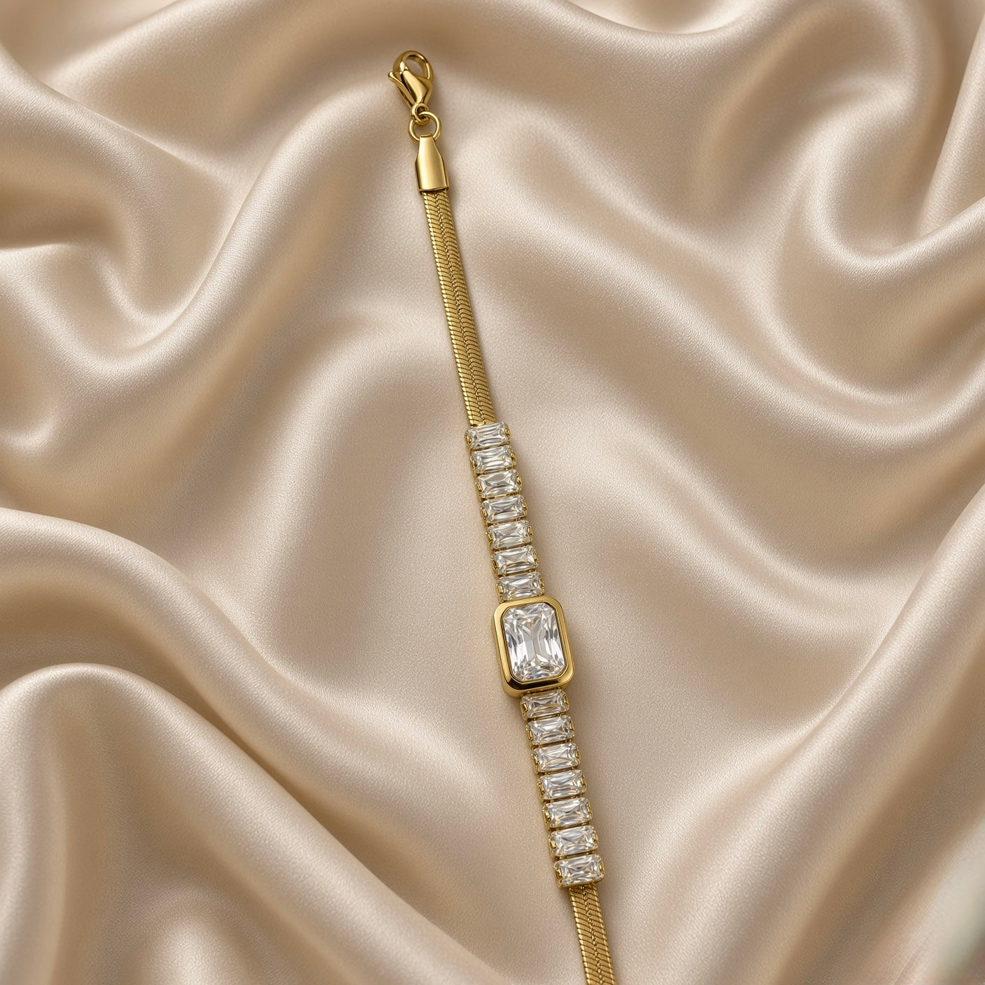 Gold Herringbone Chain Bracelet Baguette Crystal Station Emerald Bezel Centre - close detail flat lay on cream satin: angled detail view of baguette station row and emerald bezel centre showing individual channel-set baguette stone setting detail, large emerald-cut centre stone in warm gold bezel, flat herringbone chain below, cream satin background