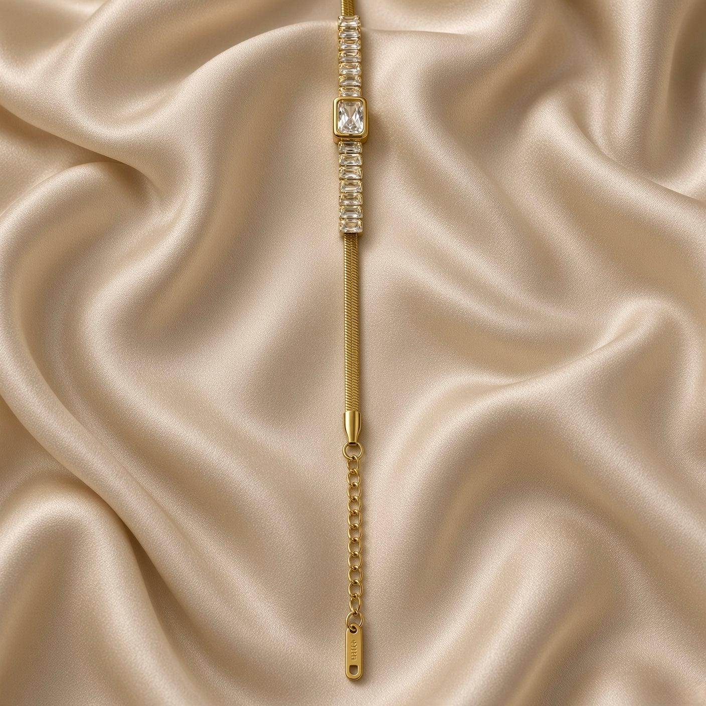 Gold Herringbone Chain Bracelet Baguette Crystal Station Emerald Bezel Centre - on wrist lifestyle: bracelet worn on elegant wrist with white satin sleeve, flat warm gold herringbone chain sitting flush against wrist, continuous baguette crystal station row and large emerald bezel centre glowing as geometric white crystal line across wrist front, lobster clasp and curb link extension chain at side, white satin background