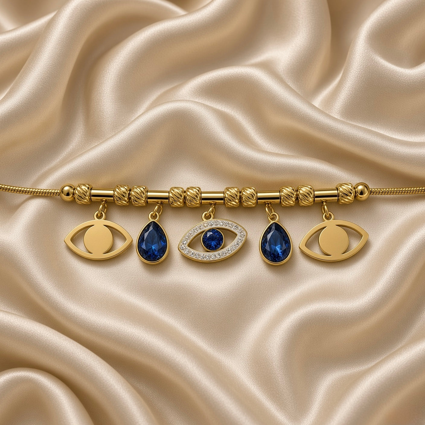 Gold Evil Eye Charm Bracelet Blue Crystal Teardrop Pavé Nazar Snake Chain - hero overhead flat lay on cream satin: full bracelet in circular form on snake chain with bead stations, 5 charms displayed: 2 open plain gold eye charms at outer positions, 2 deep sapphire blue pear-cut teardrop crystal charms in warm gold bezel, 1 central large pavé white crystal marquise evil eye charm with blue round crystal centre, cream satin background