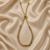 Gold Multi-Colour Emerald Cut Crystal Tennis Bracelet Bolo Adjustable Slider - overhead angled flat lay on cream satin: full bracelet showing complete multi-colour baguette crystal tennis line in circular form with warm gold box settings, bolo slider and two warm gold ball-tip chain tails visible at top, four-colour cycling crystal palette visible, cream satin background