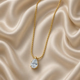 Gold Pear Cut Crystal Solitaire Pendant Necklace Three Claw Fine Cable Chain - hero portrait flat lay on cream satin: pendant necklace with fine warm 18K gold cable chain in inverted V drape on cream satin, large pear-cut white crystal zirconia solitaire in three-claw warm gold prong setting with smooth warm gold bail at centre bottom, cream satin background
