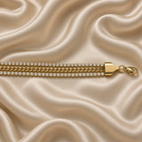 Gold Cuban Link Chain Bracelet Double Crystal Tennis Row Three Row Shield Clasp - angled overhead flat lay on cream satin: full bracelet showing complete three-row Cuban-centre crystal-flanked architecture at angle, flat-polished warm gold Cuban link chain centre between two white crystal tennis rows, cream satin background
