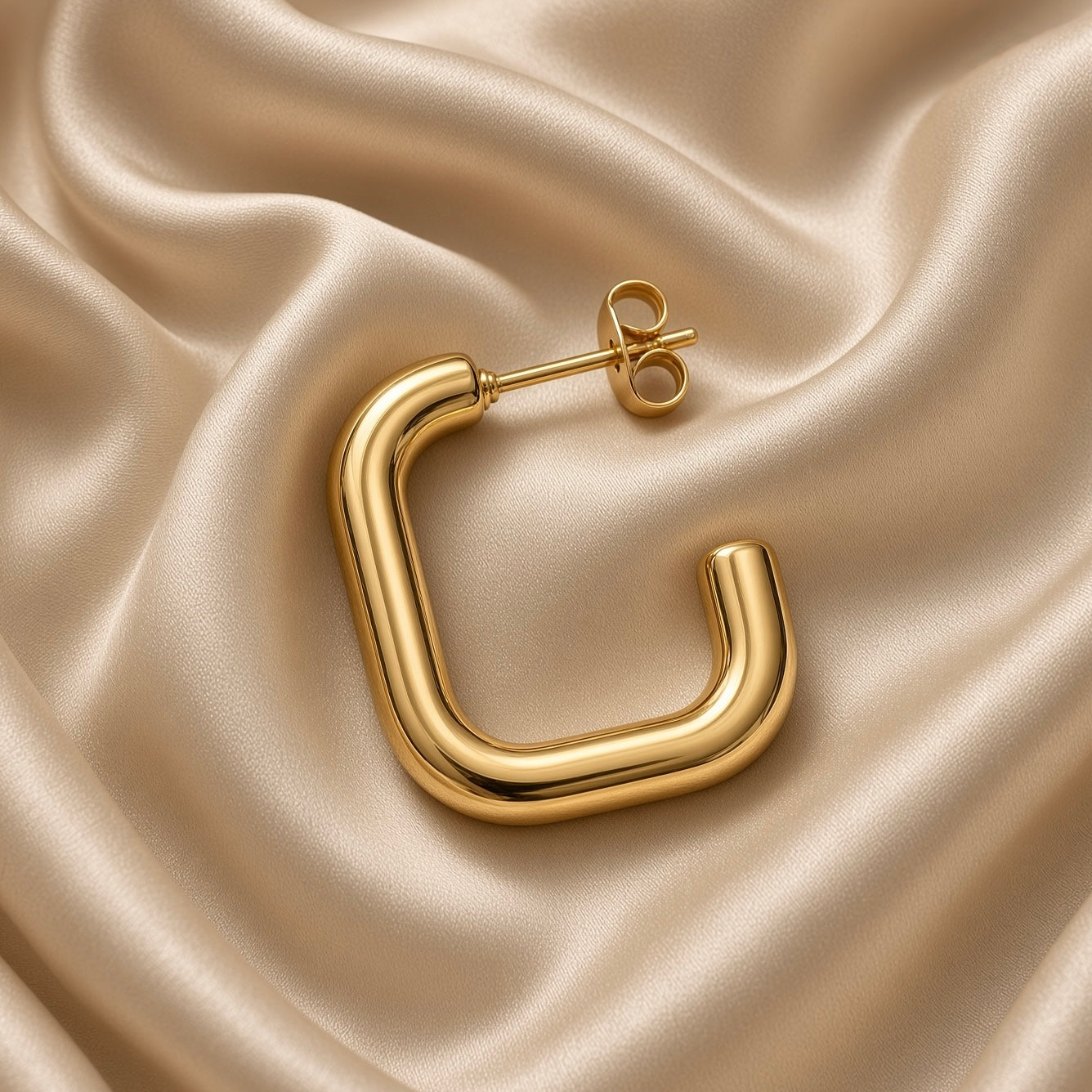 Close-up of one polished gold-tone square U-shaped hoop earring and butterfly back on champagne satin
